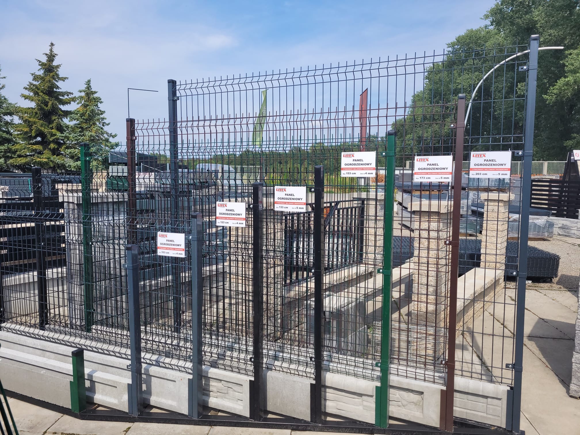 Ekspozycja różnych kolorów i rozmiarów paneli ogrodzeniowych z siatki z dołączonymi tabliczkami informacyjnymi w punkcie sprzedaży.