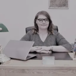 Kobieta w okularach siedzi za biurkiem w gabinecie, z laptopem, lampą, figurką Temidy i szafką z dokumentami w tle.