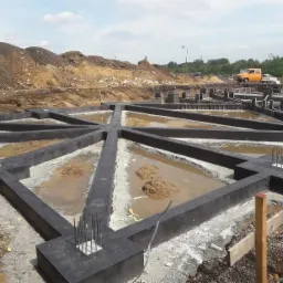 Wykopane fundamenty domu z widocznymi zbrojeniami, częściowo zalane wodą, na tle hałdy ziemi i pomarańczowej ciężarówki.