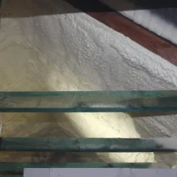 Poddasze ocieplone pianką, widoczne krokwie z zieloną impregnacją, nierównomierna struktura piany z jaśniejszymi i ciemniejszymi obszarami.