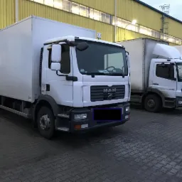 Dwa białe samochody ciężarowe typu furgon zaparkowane na brukowanym placu przed żółtym budynkiem magazynowym z oknami.