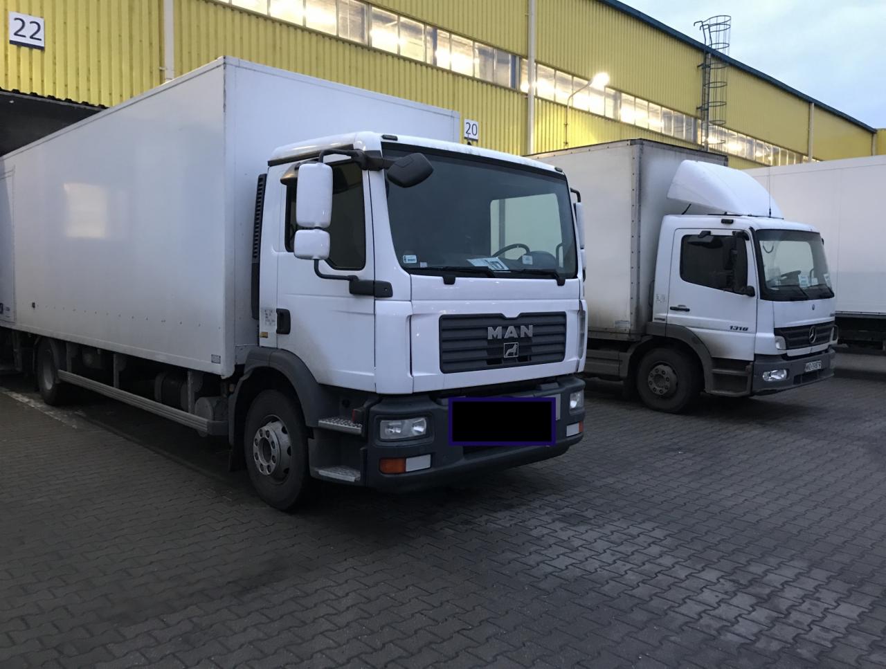 Dwa białe samochody ciężarowe typu furgon zaparkowane na brukowanym placu przed żółtym budynkiem magazynowym z oknami.