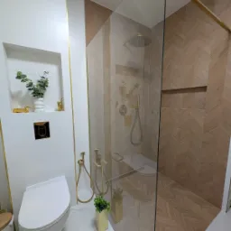 Elegancka łazienka po remoncie: kabina prysznicowa ze złotymi detalami, białe płytki z wnęką dekoracyjną i toaleta podwieszana. Nowoczesny design.