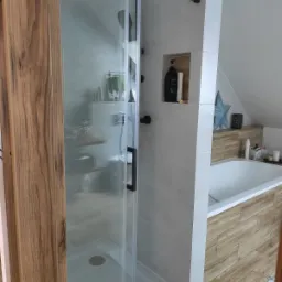 Nowoczesna łazienka na poddaszu z kabiną prysznicową typu walk-in z czarnymi detalami, białą wanną obudowaną drewnopodobnymi płytkami i wnęką w ścianie z kosmetykami.
