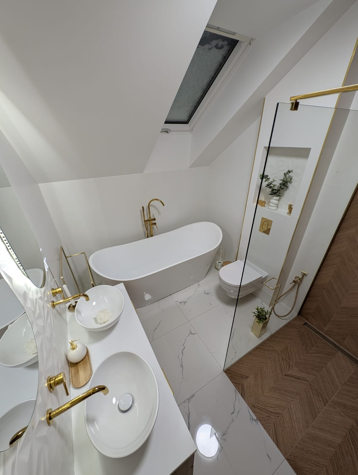Nowoczesna łazienka na poddaszu z białą armaturą i złotymi detalami. Uwagę zwraca wanna wolnostojąca, umywalki nablatowe i przeszklona kabina prysznicowa.