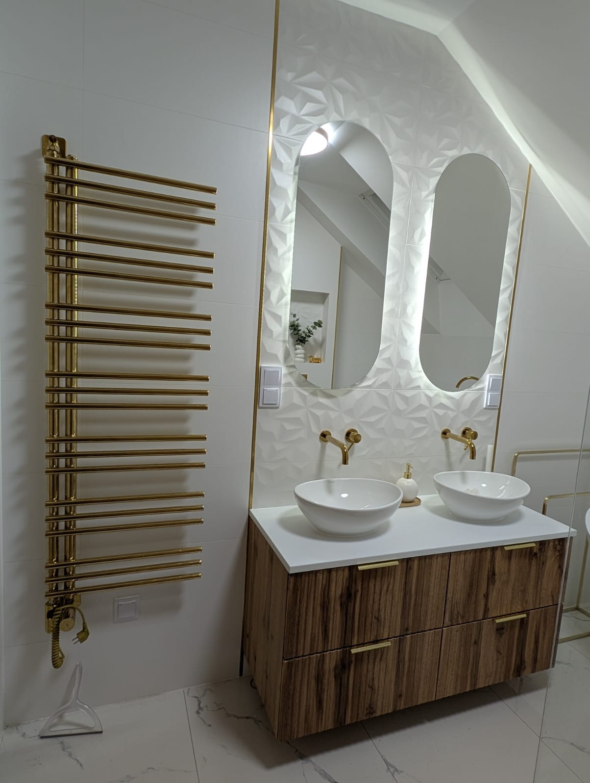 Nowoczesna łazienka na poddaszu z umywalkami nablatowymi, złotą armaturą i grzejnikiem. Białe płytki 3D i lustra z podświetleniem LED dodają elegancji.