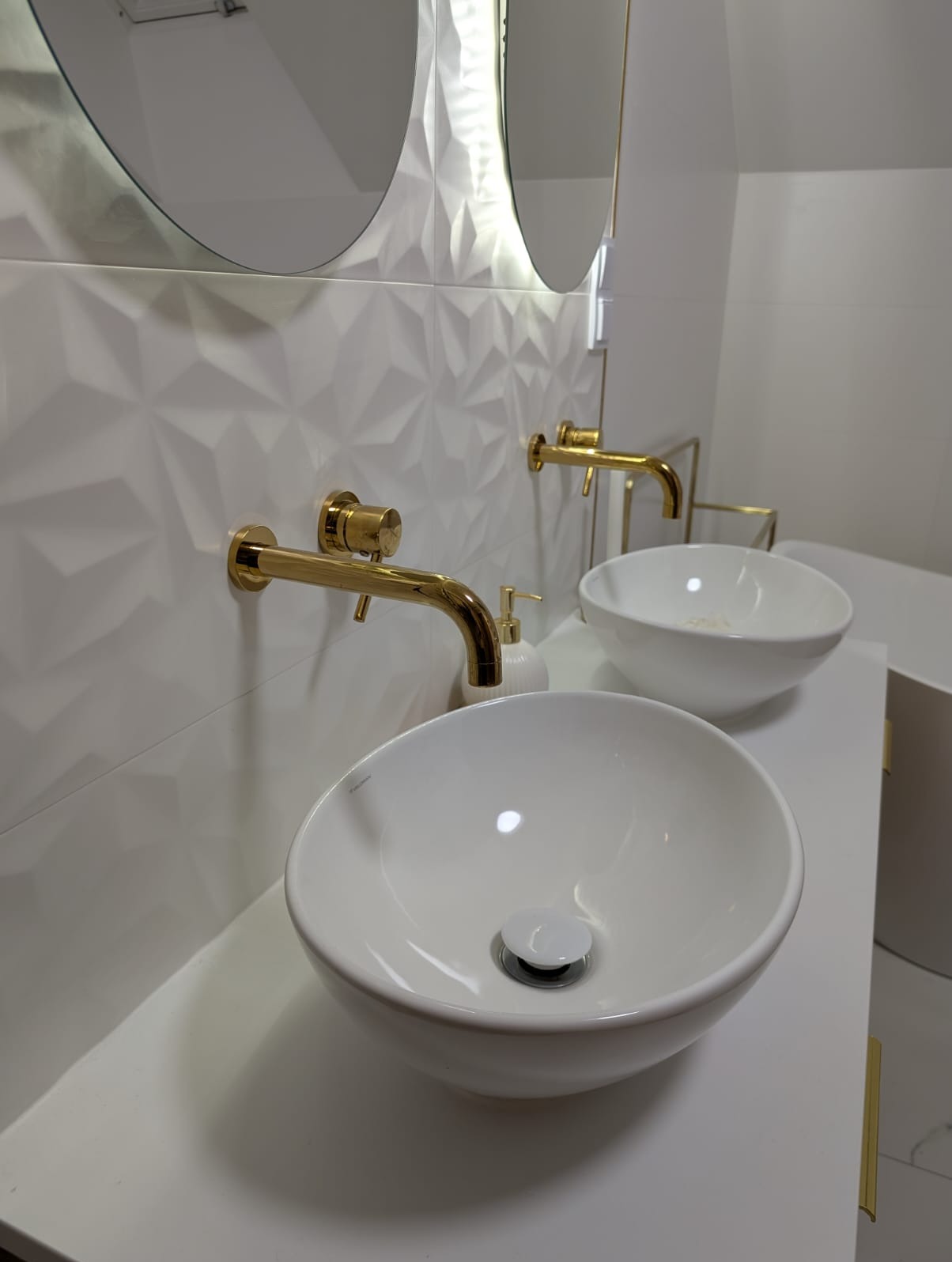 Biała łazienka z dwiema okrągłymi umywalkami na blacie, złote baterie ścienne, dekoracyjne białe płytki 3D i podświetlane lustra. Minimalistyczny design.