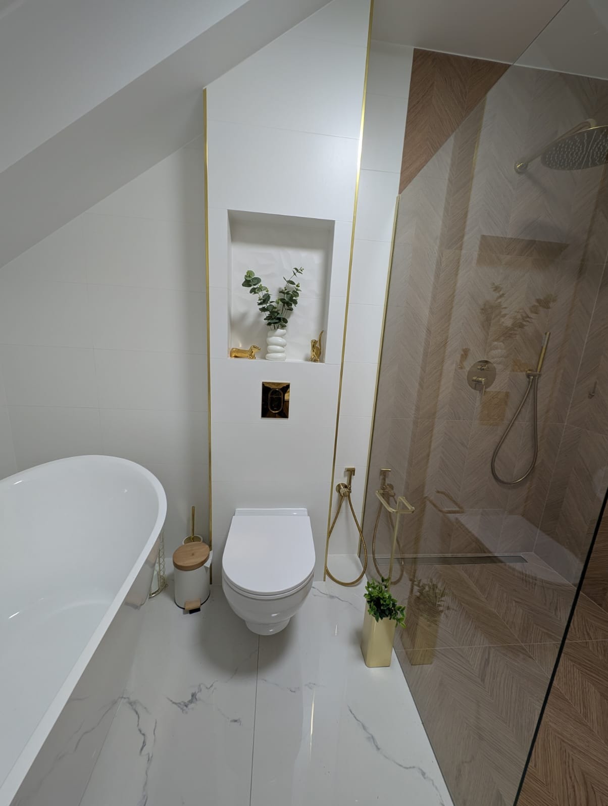 Elegancka łazienka na poddaszu z białą armaturą, złotymi akcentami i kabiną prysznicową z wzorem jodełki. Wnęka z dekoracjami. Marmurowe płytki podłogowe.