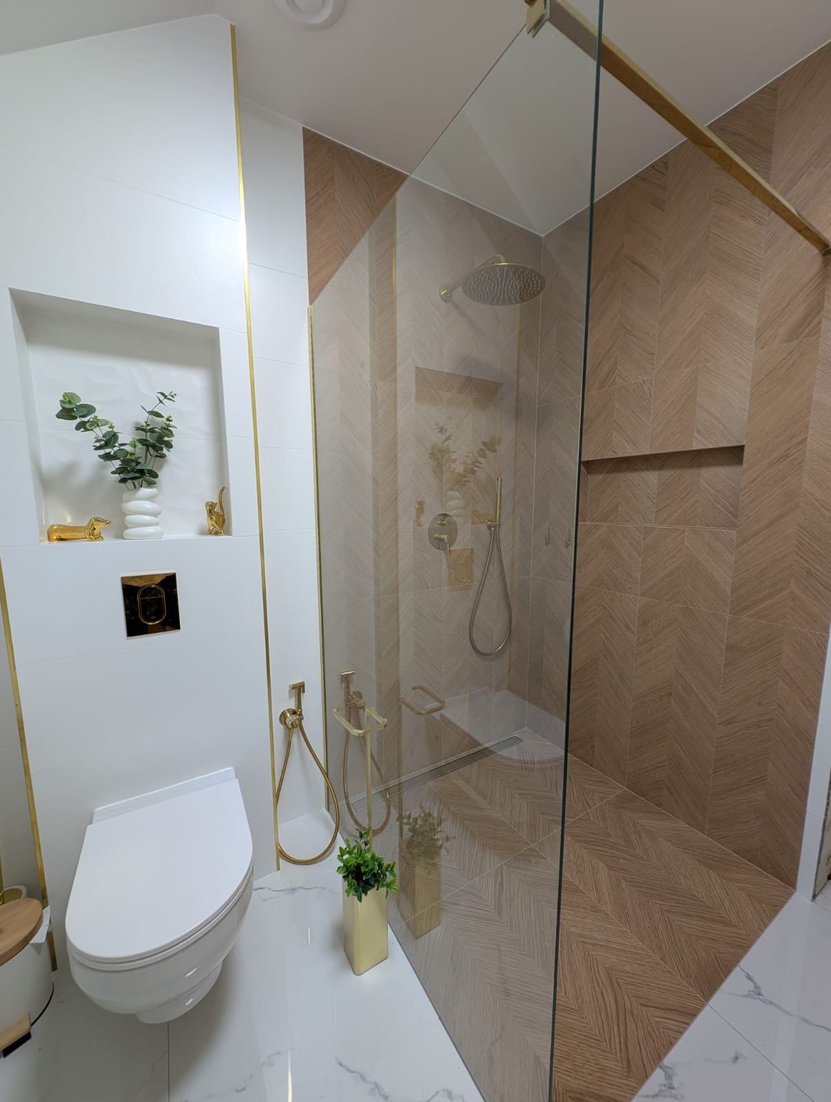 Elegancka łazienka po remoncie: kabina prysznicowa ze złotymi detalami, białe płytki z wnęką dekoracyjną i toaleta podwieszana. Nowoczesny design.