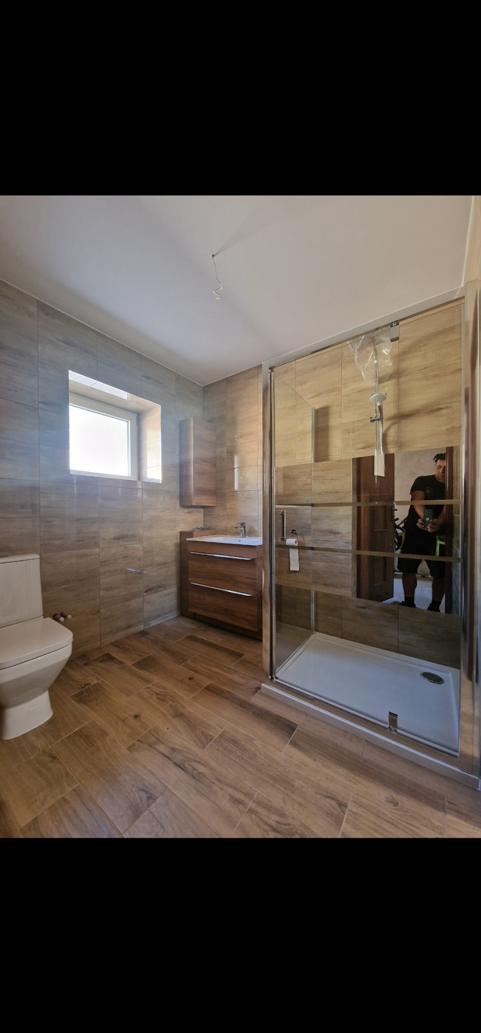 Nowoczesna łazienka z drewnopodobnymi płytkami, kabiną prysznicową ze szkła hartowanego i wiszącą szafką z umywalką. W lustrze odbija się osoba robiąca zdjęcie.