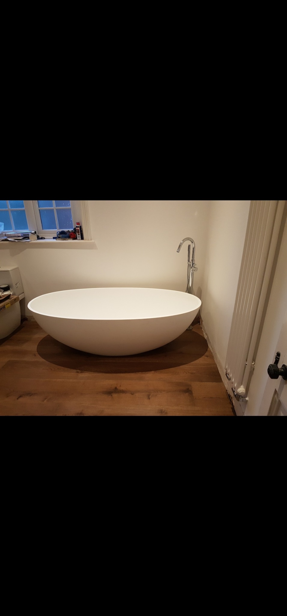 Nowoczesna, wolnostojąca wanna w kształcie misy z chromowaną baterią, umieszczona na drewnianej podłodze w jasnej łazience z grzejnikiem żeberkowym i oknem.