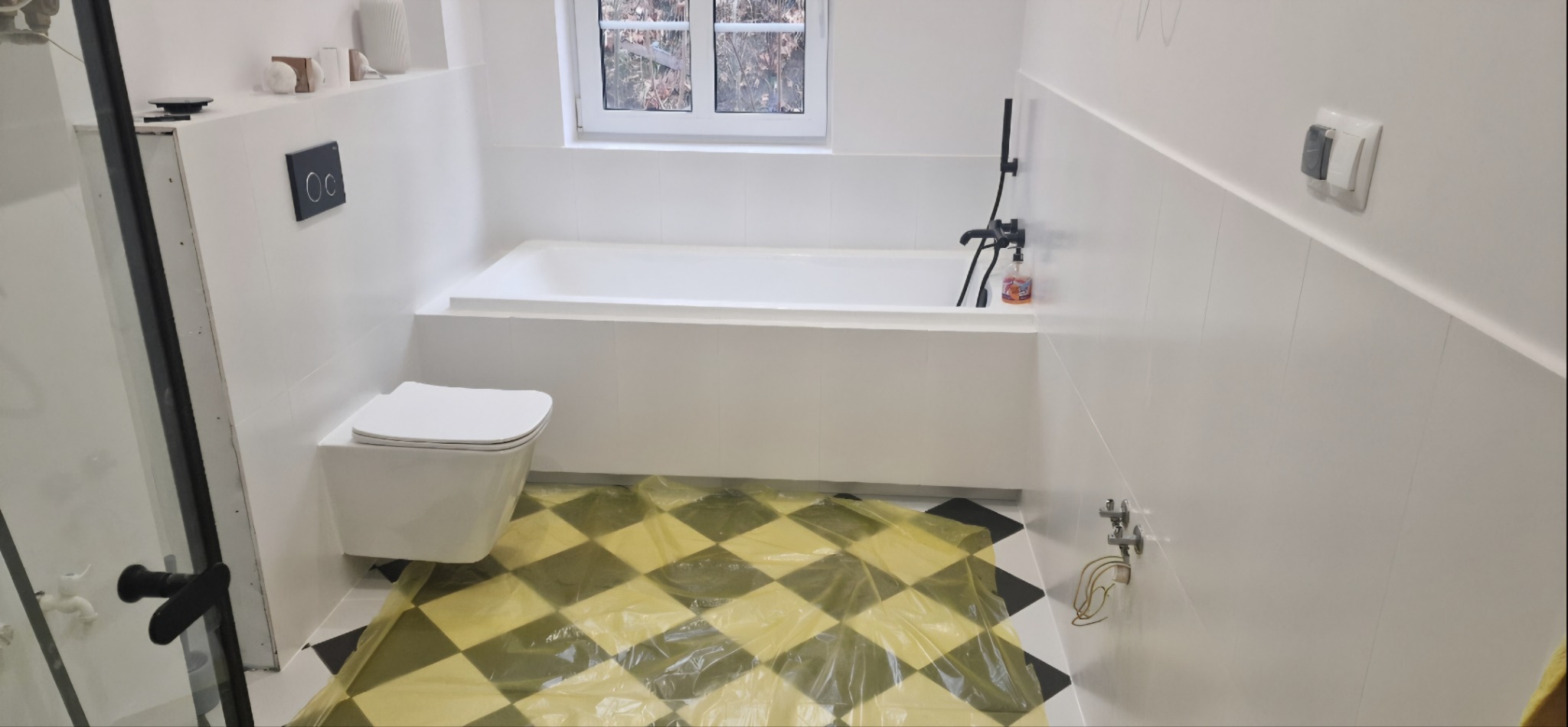 Biała łazienka z wanną i podwieszaną toaletą. Na podłodze folia ochronna z wzorem szachownicy. Minimalistyczny styl, armatura w czarnym macie.