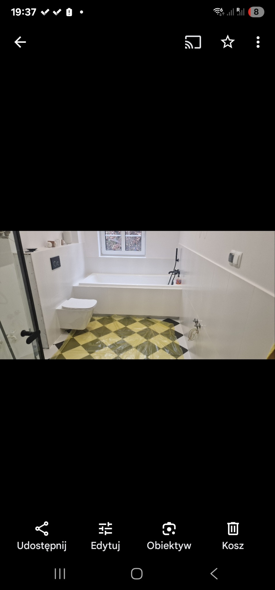 Biała łazienka po remoncie z wanną, toaletą i czarno-białą podłogą zabezpieczoną folią ochronną. Widok na okno.