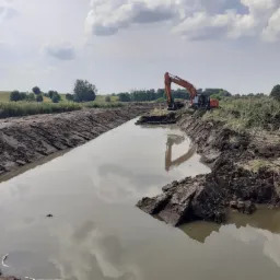 Pomarańczowa koparka Hitachi na brzegu nowo wykopanego, wypełnionego wodą stawu, odbicie chmur w wodzie, świeżo naruszona ziemia.
