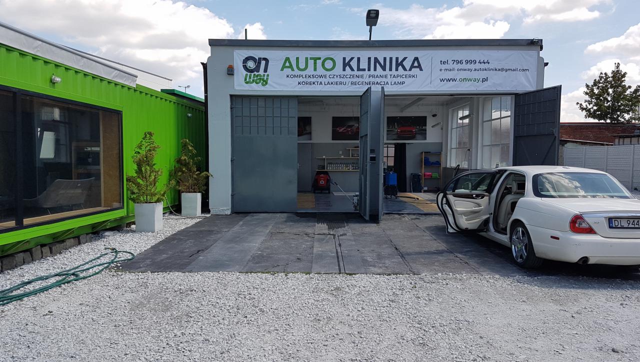 Widok na Auto Klinikę z otwartymi wrotami garażowymi, wewnątrz widoczne wyposażenie, przed budynkiem biały Jaguar z otwartymi drzwiami pasażera, obok zielony kontener biurowy i żwirowe podłoże.