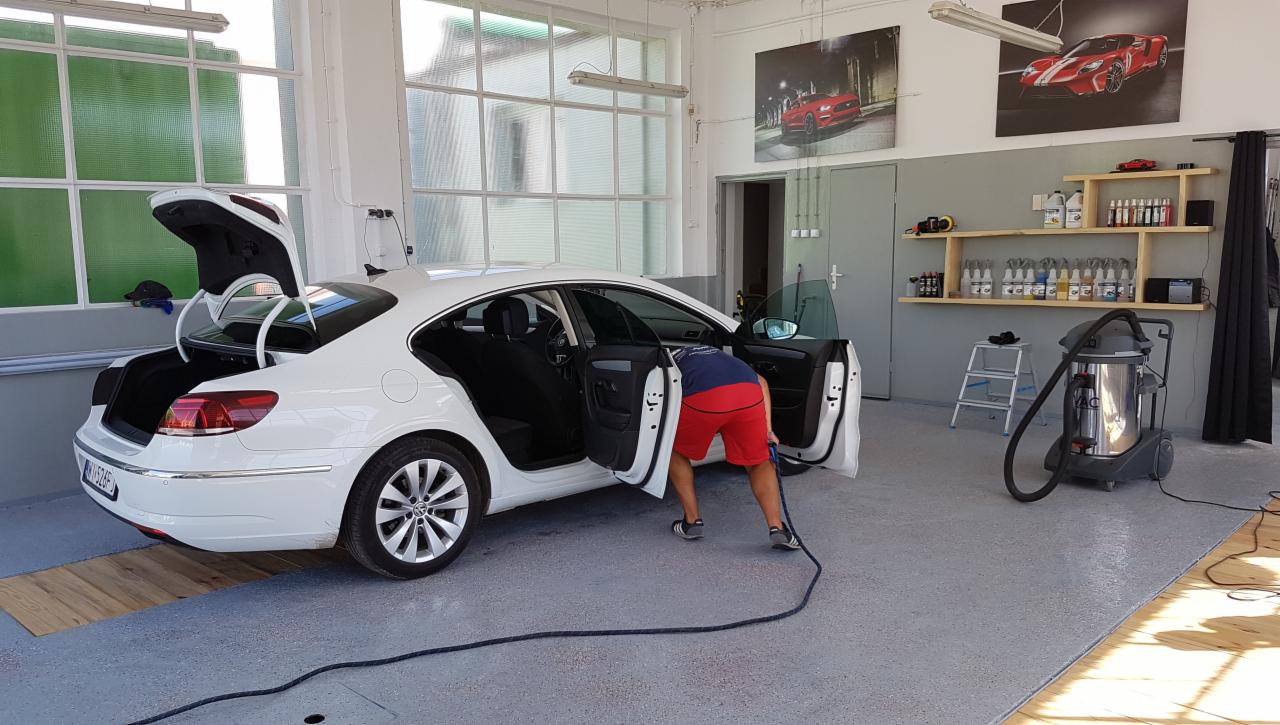 Wnętrze białego samochodu czyszczone odkurzaczem przemysłowym w warsztacie, widoczne otwarte drzwi i bagażnik, na ścianach plakaty samochodów sportowych i półki z preparatami do czyszczenia.