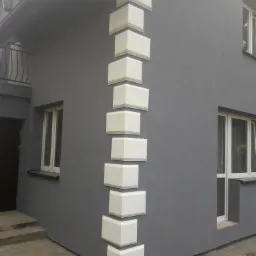 Szary budynek z narożnymi, białymi dekoracjami elewacyjnymi ułożonymi pionowo, widoczne okna i fragment balkonu z metalową balustradą.