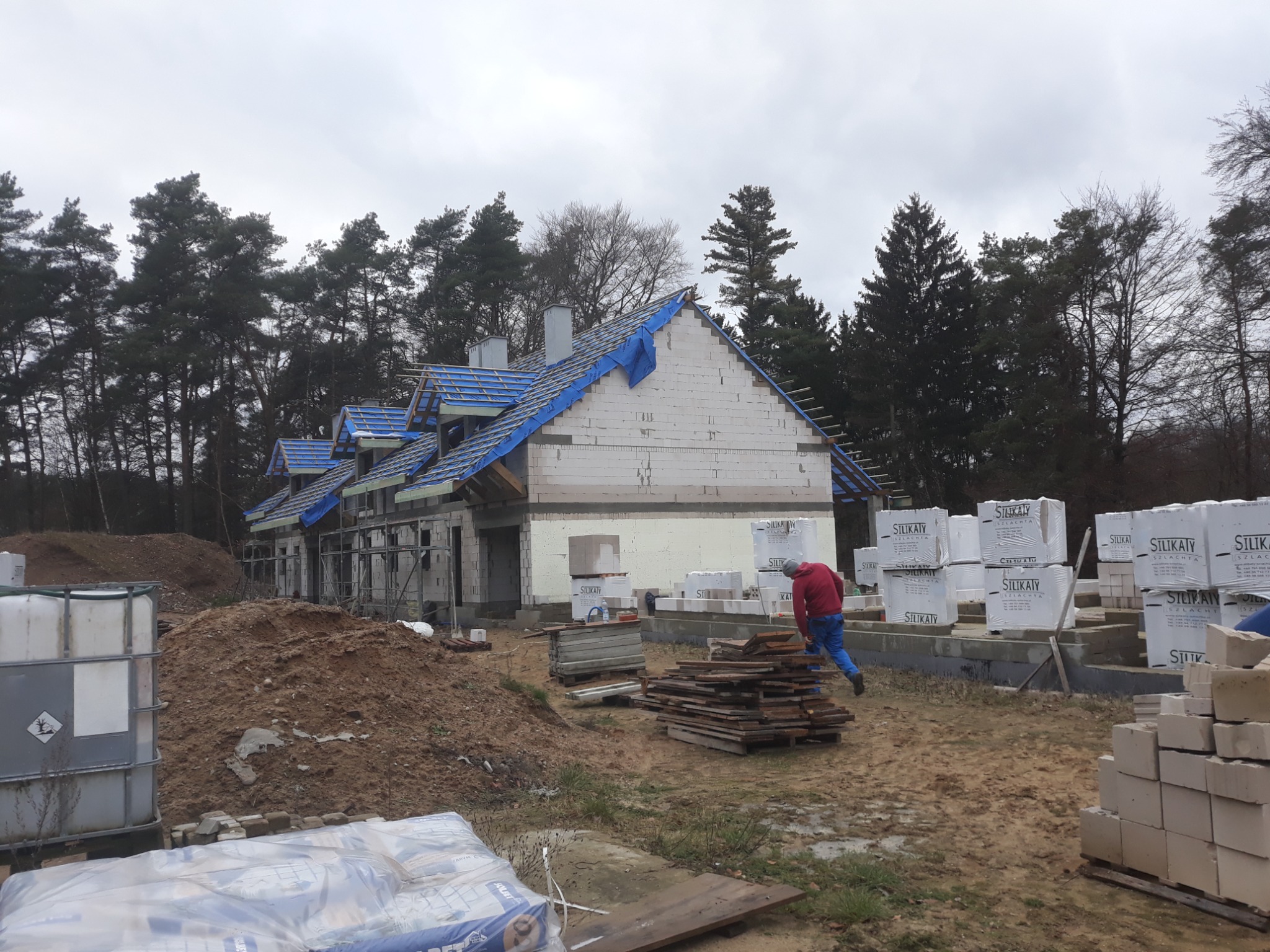 Budowa domu jednorodzinnego z białych bloczków silikatowych, widoczne ściany w stanie surowym otwartym, więźba dachowa zabezpieczona folią, stosy materiałów budowlanych oraz pracownik w czerwonej...