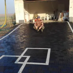 Ułożona czarna kostka brukowa z białymi liniami tworzącymi wz&oacute;r przed garażem, mężczyzna w kucającej pozycji na kostce.