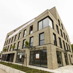 Nowoczesny budynek biurowy z cegły klinkierowej, duże okna z czarnymi ramami, widoczny napis 'Do wynajęcia' na szybie, zieleń wokół budynku