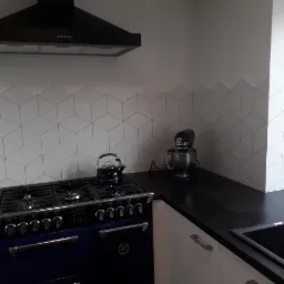 Nowoczesna kuchnia z biało-czarną kolorystyką, wyróżniająca się geometrycznymi płytkami ściennymi tworzącymi iluzję trójwymiarowości, ciemnym blatem i niebieską kuchenką retro.