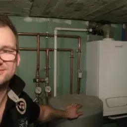Uśmiechnięty mężczyzna w okularach prezentuje kotłownię z białym piecem grzewczym i miedzianą instalacją, widoczne manometry i zbiornik akumulacyjny.