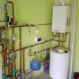 Kompletna instalacja grzewcza w pomieszczeniu z piecem gazowym, zbiornikiem akumulacyjnym i rozbudowanym systemem rur miedzianych z manometrami i pompami obiegowymi na ścianie o pistacjowym odcieniu.