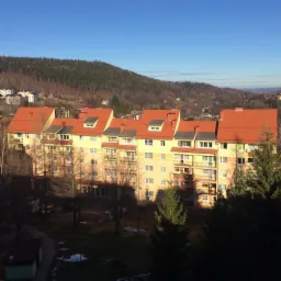 Wielopiętrowy budynek mieszkalny z pomarańczowym dachem i szarymi obróbkami blacharskimi, widok z góry na tle zalesionych wzgórz i błękitnego nieba.