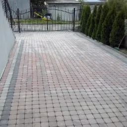 Ułożona kostka brukowa w kolorze beżowo-szarym tworząca wzór na podjeździe, z widocznym fragmentem metalowej bramy i rzędem tuj w tle.