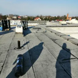Widok z góry na płaski dach pokryty papą termozgrzewalną, z rolkami materiału na palecie i pojedynczą rolką leżącą na dachu; w tle zabudowania miejskie i kościół.