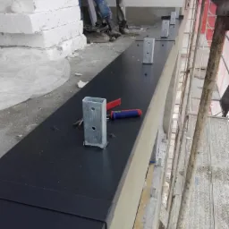 Montaż obróbki blacharskiej na balkonie, widoczne metalowe elementy konstrukcyjne, narzędzia i robotnicy w tle.