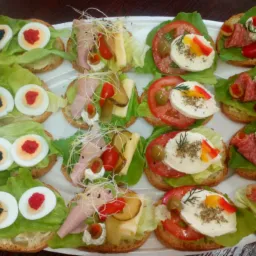 Taca z różnorodnymi kanapkami na jasnym pieczywie: jedne z jajkiem przepiórczym barwionym na czarno i czerwono, inne z wędliną, serem, warzywami i kiełkami, ułożone dekoracyjnie na liściach sałaty.