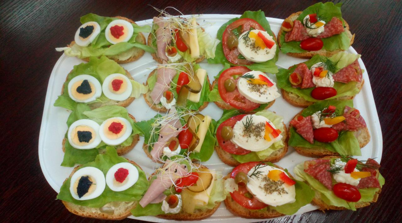 Taca z różnorodnymi kanapkami na jasnym pieczywie: jedne z jajkiem przepiórczym barwionym na czarno i czerwono, inne z wędliną, serem, warzywami i kiełkami, ułożone dekoracyjnie na liściach sałaty.