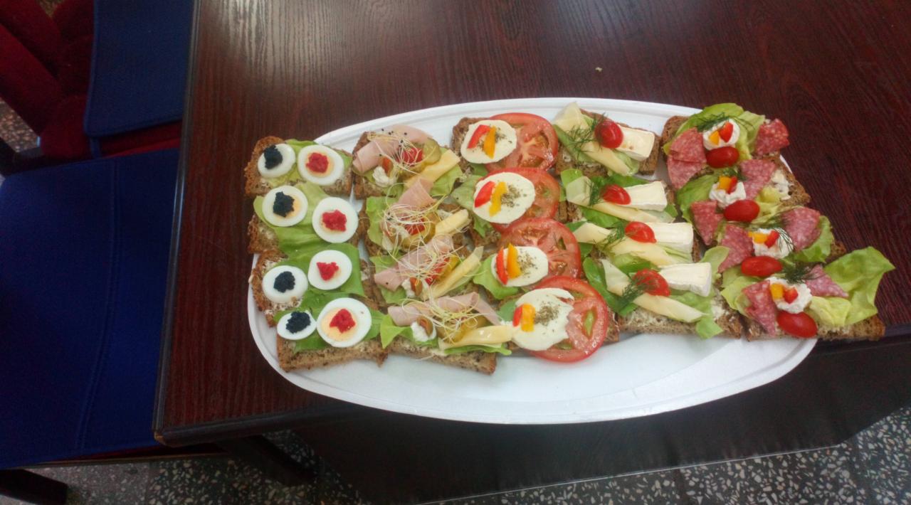 Talerz z różnorodnymi kanapkami na ciemnym pieczywie, dekorowane jajkiem, warzywami, serem i wędlinami; widoczne detale jak kiełki i pomidorki cherry.