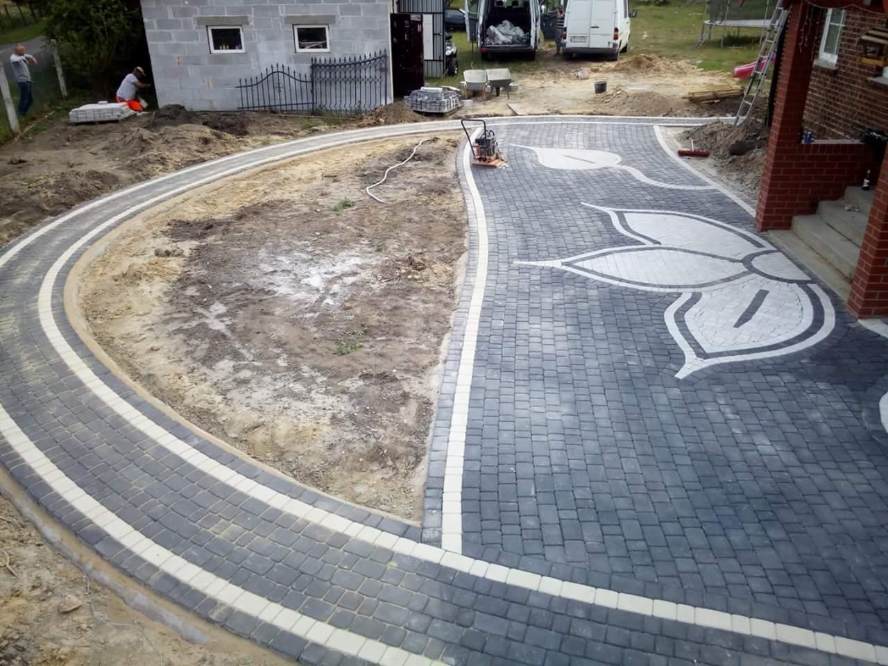 Ukończona ścieżka i podjazd z kostki brukowej w kształcie kwiatu, ciemnoszara kostka z jasnymi obrzeżami, widoczna ubijarka wibracyjna i budynek w tle.