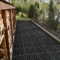 Przygotowany teren pod ścieżkę z czarną kratką drenażową, drewniany składzik na drewno po lewej stronie, widok na ogród w tle.