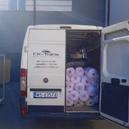 Otwarty biały bus marki Fiat, załadowany rolkami materiału, z widocznym logo firmy transportowej CK-Trans i numerem rejestracyjnym WS 6357A, na tle szarego budynku.