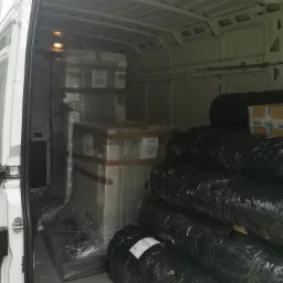 Wnętrze białego busa załadowane zapakowanymi meblami owiniętymi folią stretch oraz czarnymi workami foliowymi, widoczne etykiety 'Ostrożnie Szkło' na paczkach.