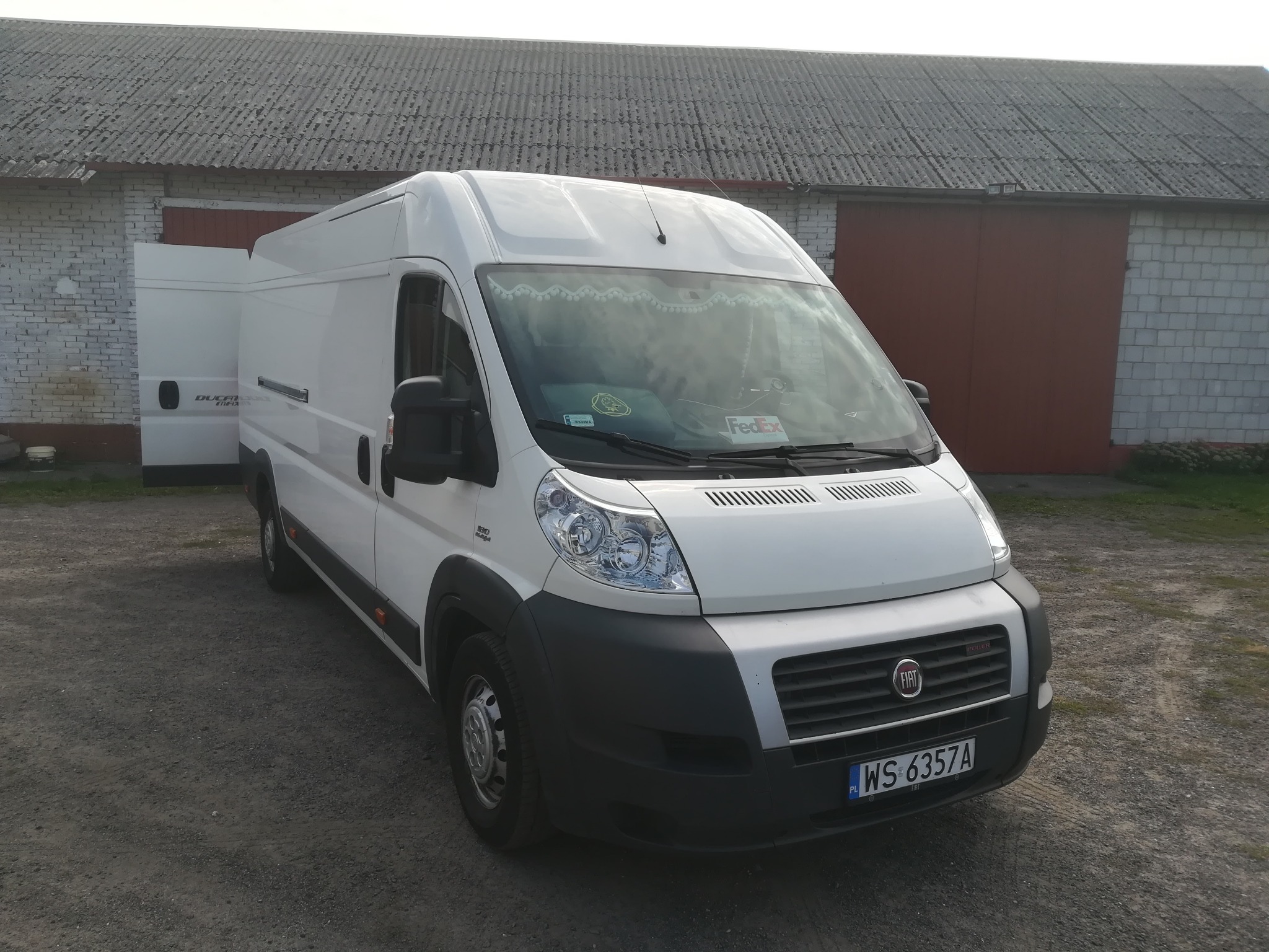 Biały bus Fiat Ducato z otwartymi drzwiami bocznymi, zaparkowany na żwirowym podłożu przed budynkiem gospodarczym z szarym dachem.
