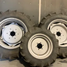 Trzy koła od ciągnika rolniczego, z widocznymi oponami o agresywnym bieżniku i jasnoszarymi felgami, ustawione na podłodze w pomieszczeniu o metalowych ścianach.