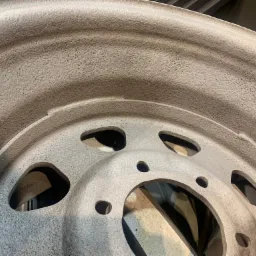 Metalowa felga samochodowa po obróbce, pokryta szorstką, jednolitą powłoką w odcieniach szarości. Widoczne otwory montażowe i wentylacyjne.