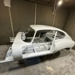 Karoseria klasycznego samochodu Jaguar E-Type po piaskowaniu i wstępnym malowaniu, w kabinie lakierniczej z widocznymi śladami po procesie.