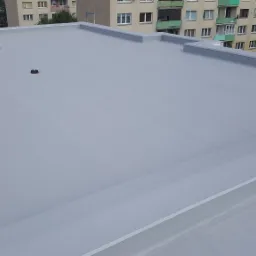 Szary, płaski dach pokryty nową membraną, z widocznymi detalami obróbki blacharskiej i elementami wentylacyjnymi na tle budynku mieszkalnego.