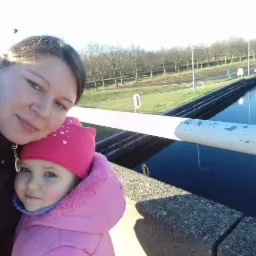 Uśmiechnięta mama z córką w różowej kurtce na tle kanału z czystą wodą i drzewami. Słoneczny dzień, świeże powietrze.