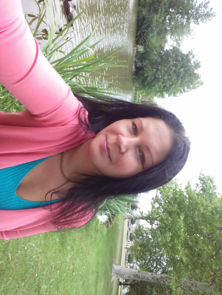 Uśmiechnięta kobieta w różowym kardiganie robi selfie na tle jeziora z kaczkami i zieleni. Zdjęcie obrócone o 90 stopni w lewo.