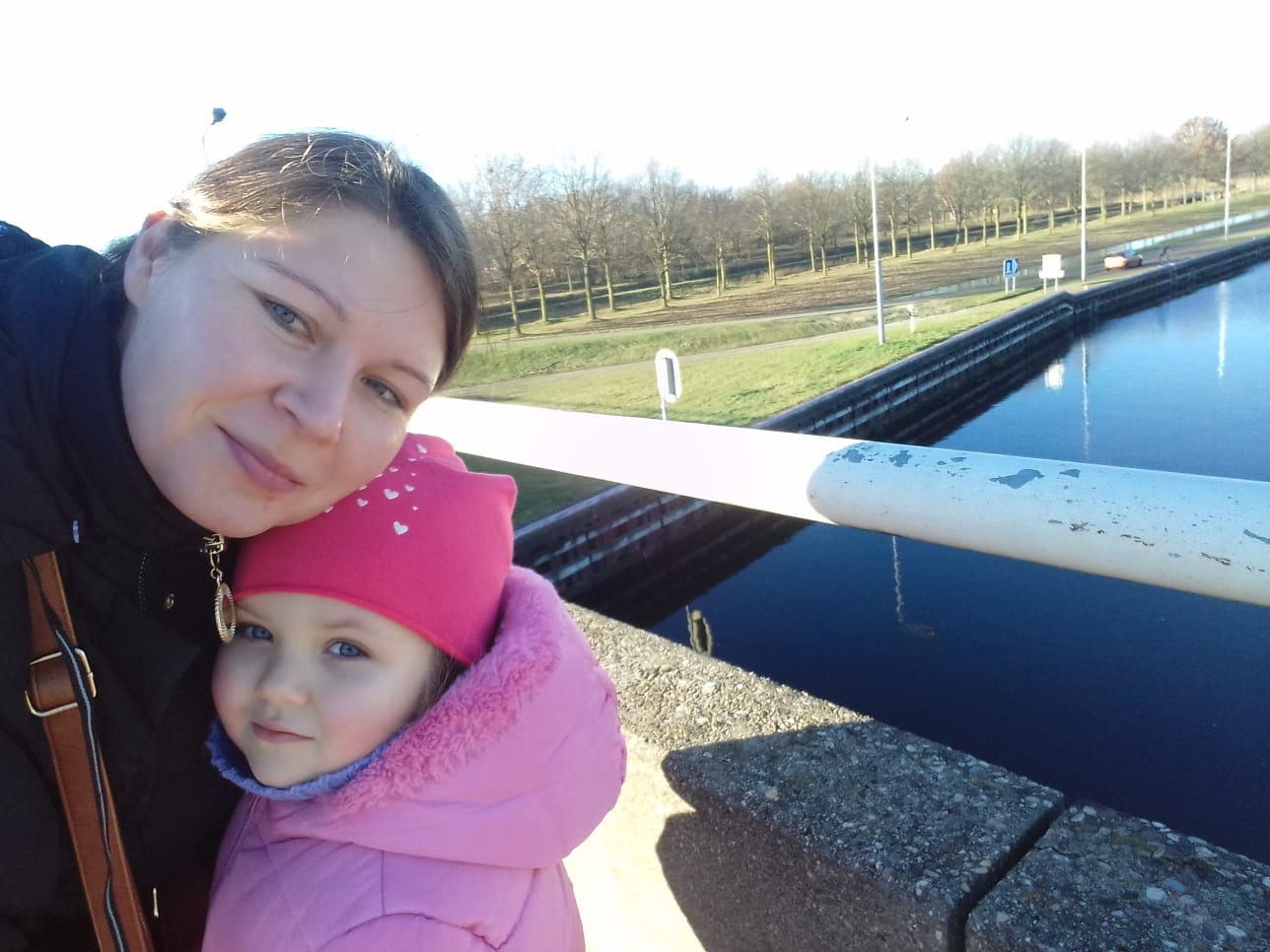 Uśmiechnięta mama z córką w różowej kurtce na tle kanału z czystą wodą i drzewami. Słoneczny dzień, świeże powietrze.