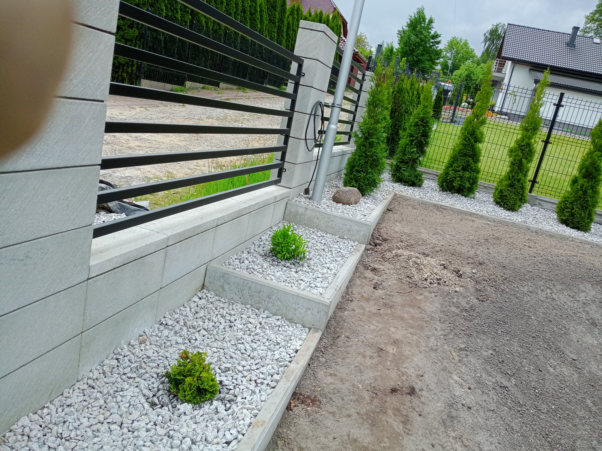 Kaskadowy ogródek skalny z jasnymi kamykami, niskie tuje w rzędzie wzdłuż ogrodzenia z metalowymi przęsłami i betonowymi słupkami.