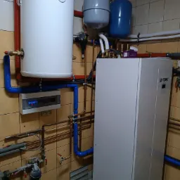 Instalacja z pompą ciepła York w kotłowni z beżowymi płytkami, widoczne zbiorniki wyrównawcze, rury miedziane i niebieskie izolacje, na podłodze wiadro.