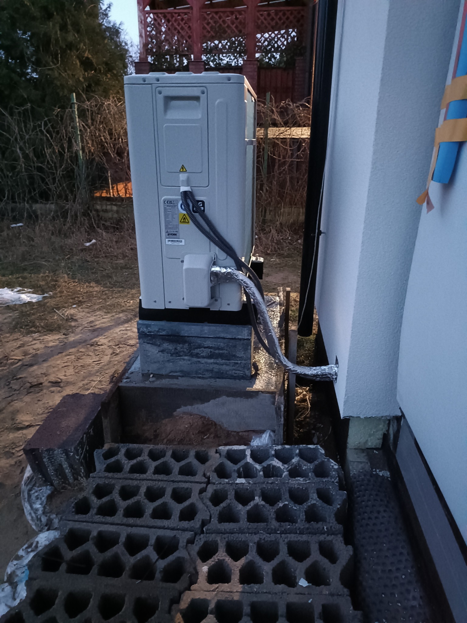 Zewnętrzna jednostka pompy ciepła zamontowana na betonowym fundamencie przy ścianie budynku, widoczne przewody w izolacji oraz stos pustaków betonowych w tle.