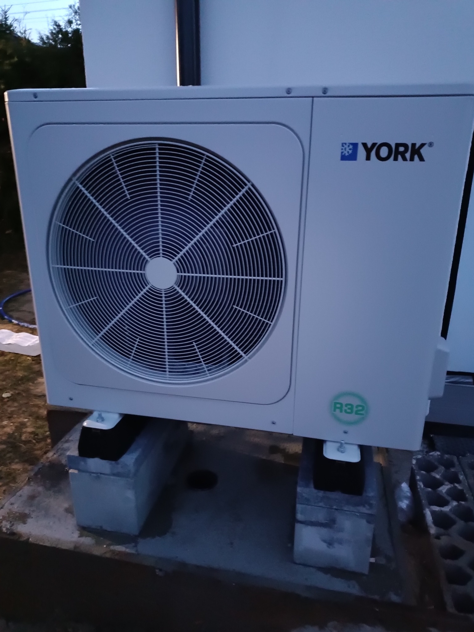 Zewnętrzna jednostka klimatyzacji York zamontowana na betonowych blokach, widoczny zielony napis R32, tło z elementami trawy i elewacji budynku.